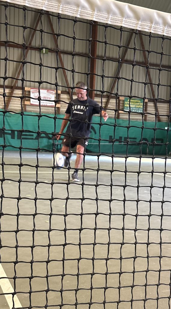 Olivier Miranda frappe au pied un ballon de rugby sur un court de tennis couvert, vu à travers le filet